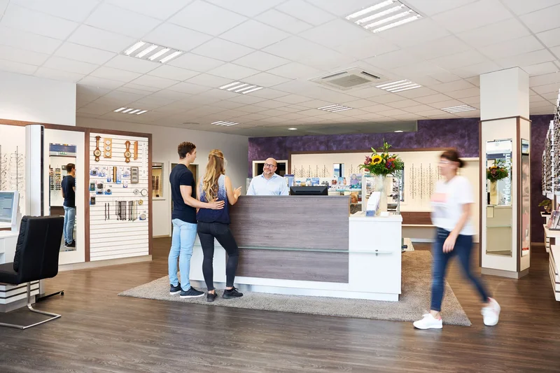 Das Bild zeigt das freundliche Ambiente eines modernen Optikgeschäfts. Ein Optiker steht hinter dem Beratungstresen und spricht mit einem jungen Paar, das Unterstützung bei der Auswahl von Brillen benötigt. Eine weitere Kundin bewegt sich durch den Rau
