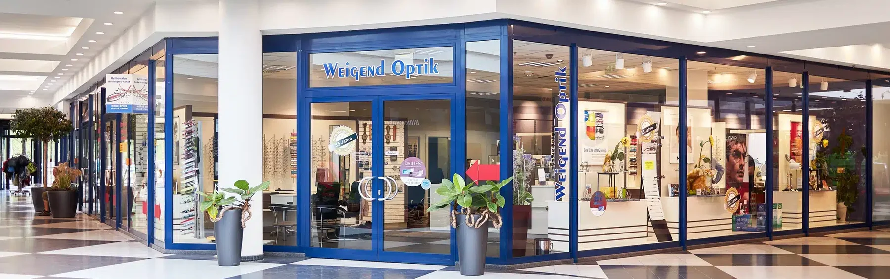 Das Bild zeigt die einladende Fassade des Optikgeschäfts „Weigend Optik“ in einem Einkaufszentrum. Die große Glasfront bietet Einblicke in das Geschäft, das mit einer modernen Einrichtung und einer breiten Auswahl an Brillen überzeugt.